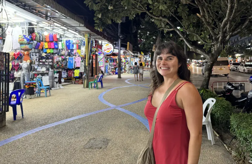 Bárbara Rocha Alcantelado sorrindo, vestida de vermelho, em uma rua movimentada de Koh Phi Phi à noite, com lojas iluminadas ao fundo