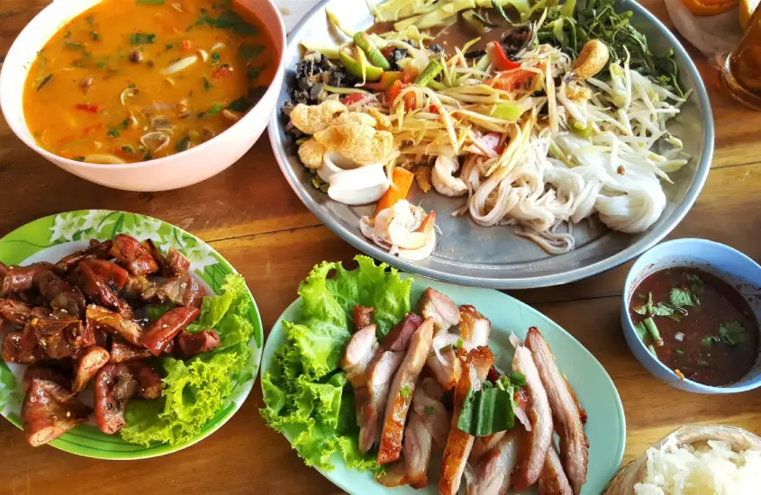 Pratos de comida tailandesa servidos em uma mesa de madeira, com sopa, carnes grelhadas e vegetais frescos