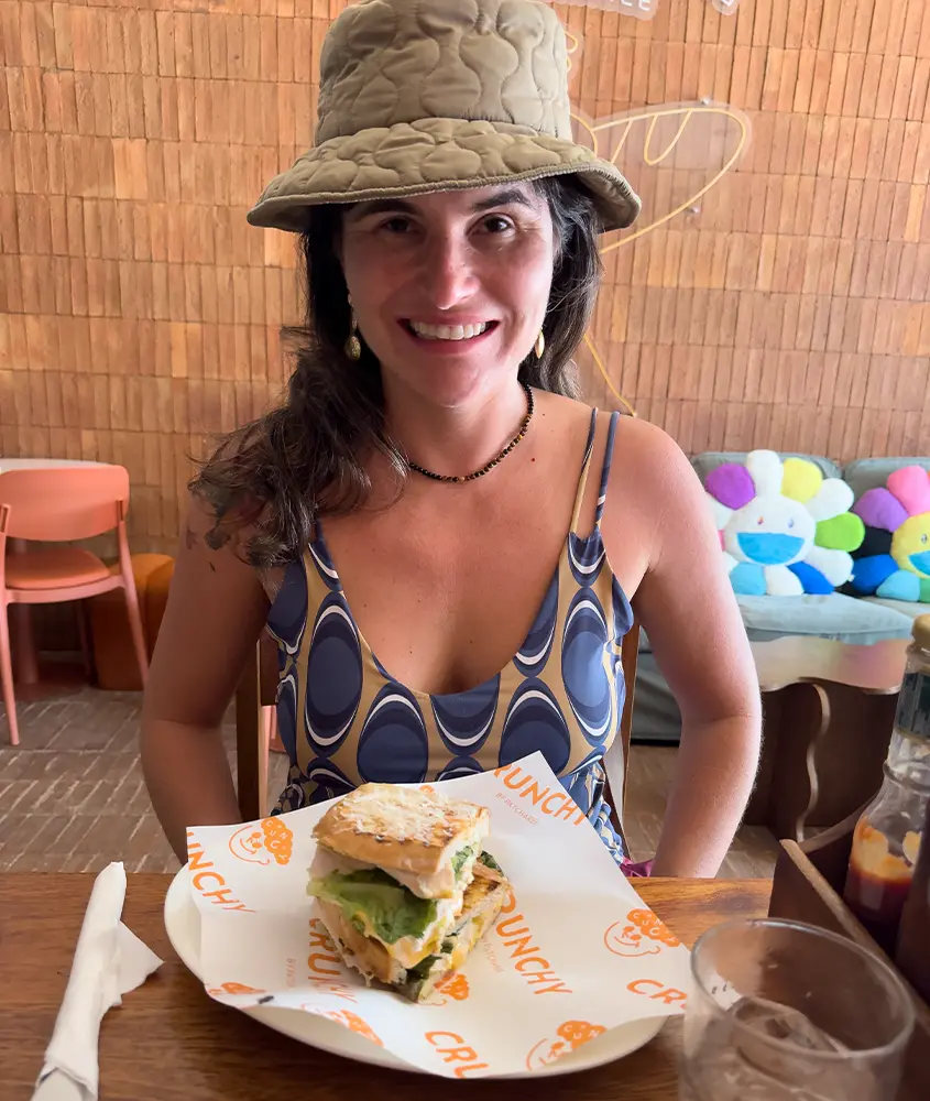 Bárbara Rocha Alcantelado usando chapéu, sentada em um restaurante em Koh Phi Phi com um sanduíche grande à sua frente.