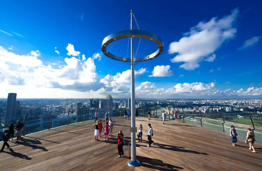 Pessoas passeando no Sands SkyPark Observation Deck, Singapura, com céu azul e vista panorâmica da cidade ao fundo.