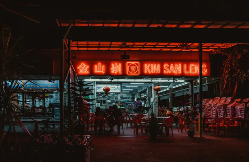 Fachada iluminada em neon vermelho do restaurante Kim San Leng, Singapura, à noite, com pessoas comendo nas mesas ao ar livre.