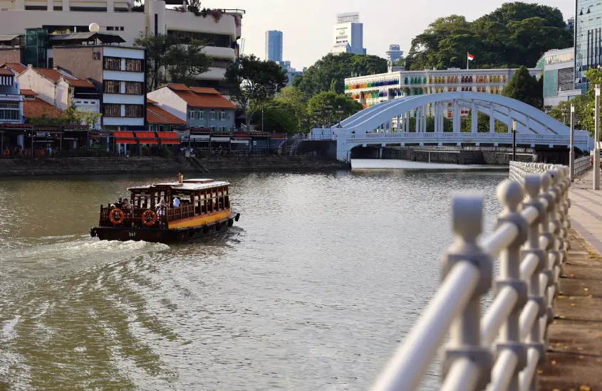 Barco tradicional navegando pelo Rio Singapura durante o dia, com uma ponte e prédios ao fundo.