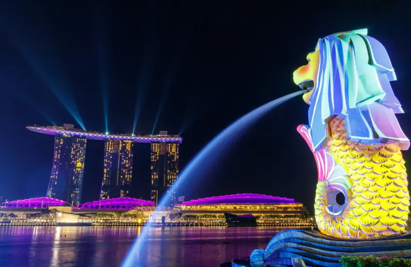 Estátua do Merlion em Singapura, iluminada à noite, com o Marina Bay Sands ao fundo também iluminado em tons de roxo