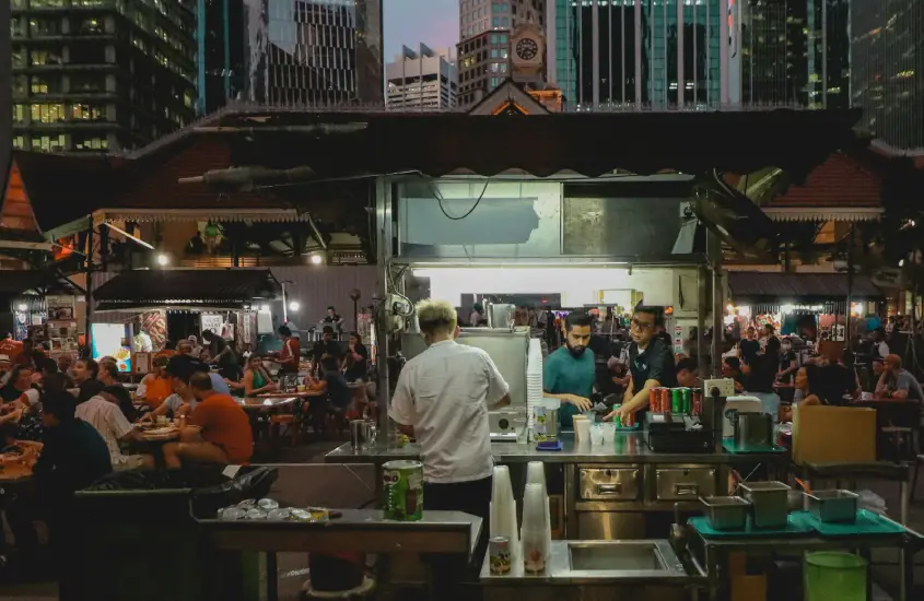 Hawker center em Singapura à noite, com pessoas comendo ao ar livre.