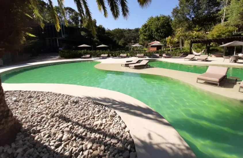 Piscina externa em forma de lago no Unique Garden, iluminada pela luz do dia, cercada por espreguiçadeiras e vegetação exuberante