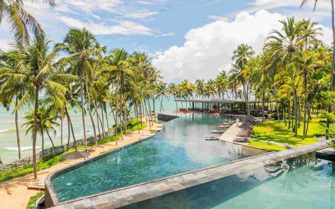 Piscina ao ar livre cercada por palmeiras e vista para o mar em resort de luxo no Brasil, durante o dia