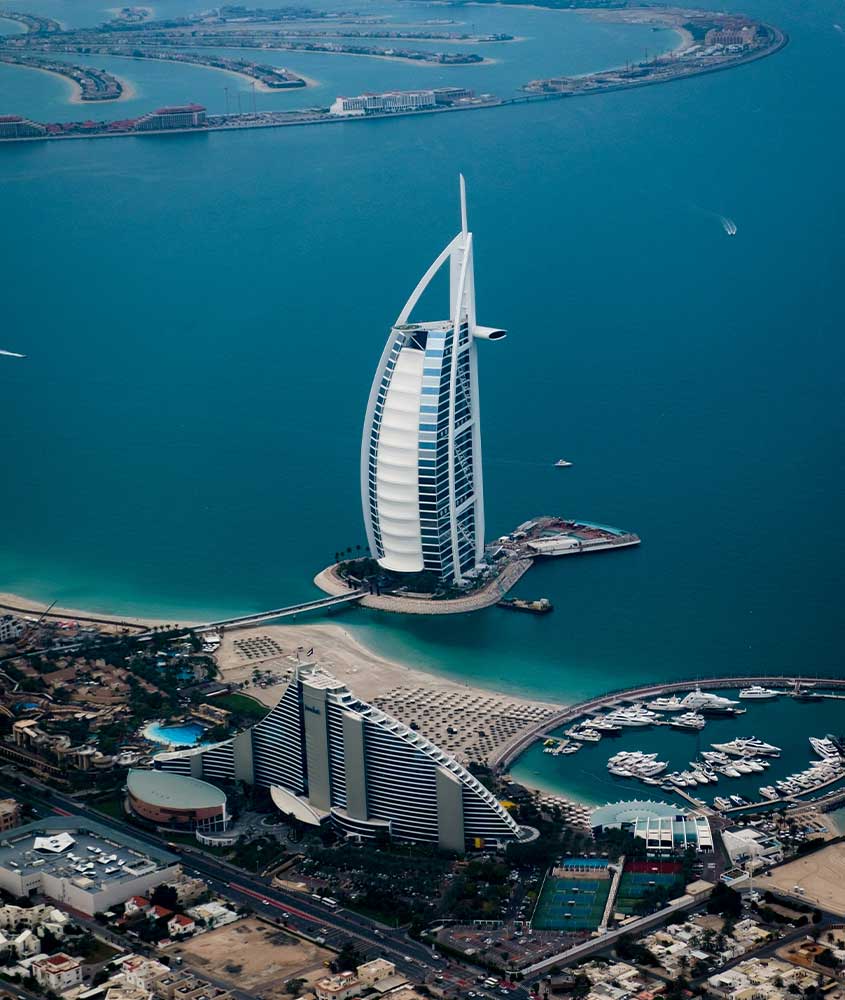 Um grande edifício em formato de vela visto de cima, com uma marina e iates ancorados, exibindo a grandiosidade da costa de Dubai e suas construções de luxo.