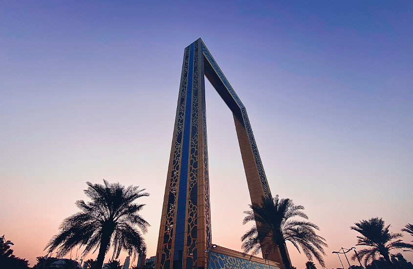 O icônico Dubai Frame ao amanhecer, com palmeiras ao redor e o céu azul-púrpura ao fundo.