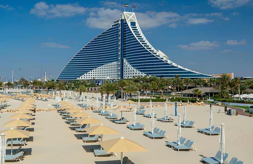 Um edifício moderno em formato de vela, destacando-se contra o céu claro, com palmeiras nas laterais. O edifício é uma das construções icônicas de Dubai.