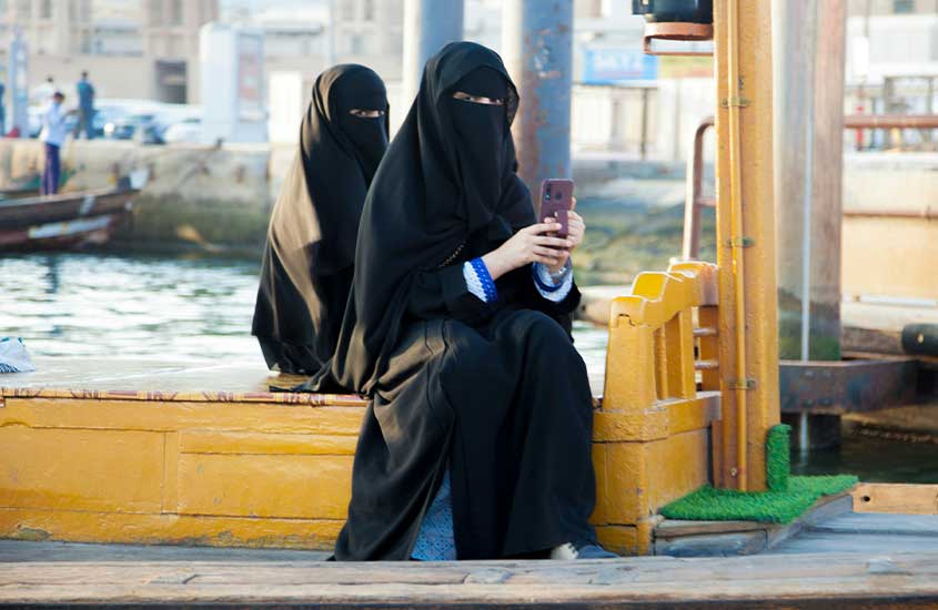 Duas mulheres vestidas com niqab, sentadas à beira de um cais. Uma delas segura um smartphone, sugerindo um momento casual e moderno, ainda que enraizado na tradição cultural.