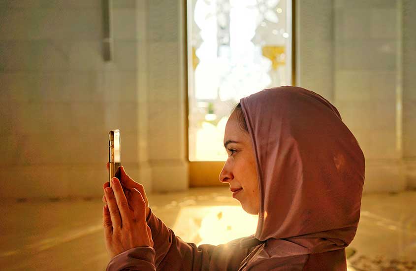 Uma mulher de hijab usa o celular para capturar a beleza de uma mesquita durante o dia.