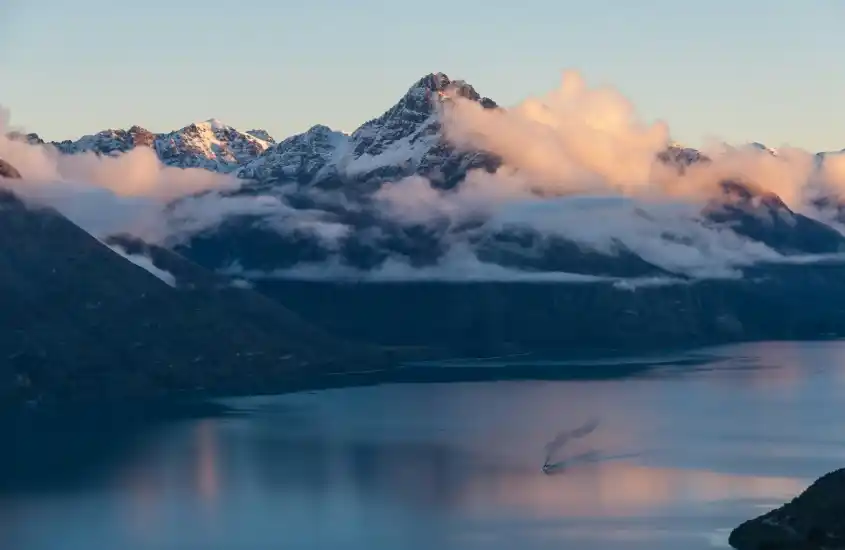 Pico montanhoso envolto em nuvens, refletido nas águas tranquilas de um lago ao amanhecer em Queenstown, um dos melhores destinos de inverno do mundo