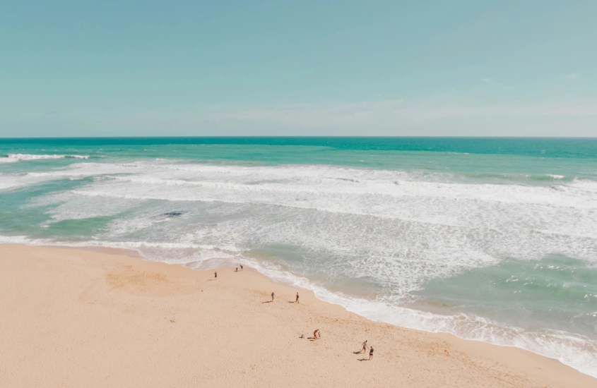 Vista aérea de uma praia deserta com areia clara e mar azul-turquesa, próximo a uma área verde densa. Beleza natural australiana