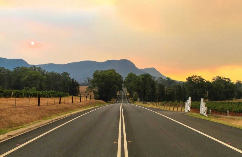 Estrada deserta entre vinhedos e montanhas, sob um céu alaranjado ao pôr do sol na Austrália. Cenário típico das regiões vinícolas