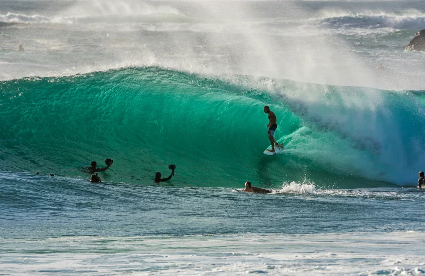 Surfista equilibrado em uma grande onda turquesa, enquanto outros observam ao redor na água