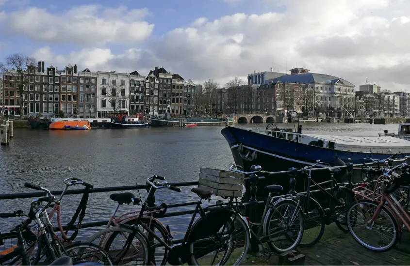 Fachada de edifícios tradicionais à beira de um canal em Amsterdã, com bicicletas estacionadas ao longo do guarda-corpo e barcos no canal