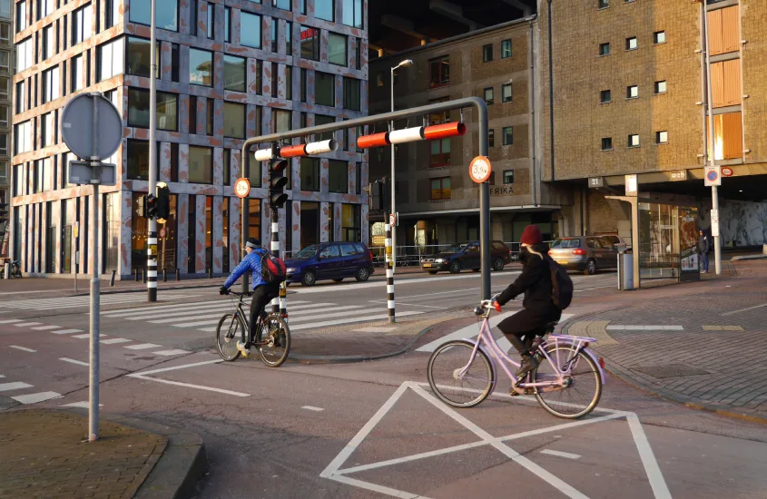 Duas pessoas andando de bicicleta enquanto cruzam uma rua em Amsterdam, cercados por prédios modernos