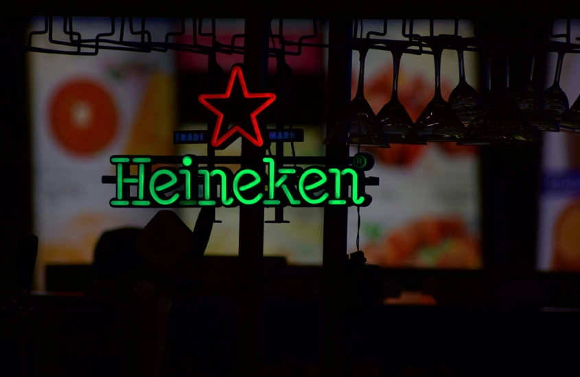 Placa de neon verde e vermelha da Heineken em um bar escuro, com copos pendurados ao fundo