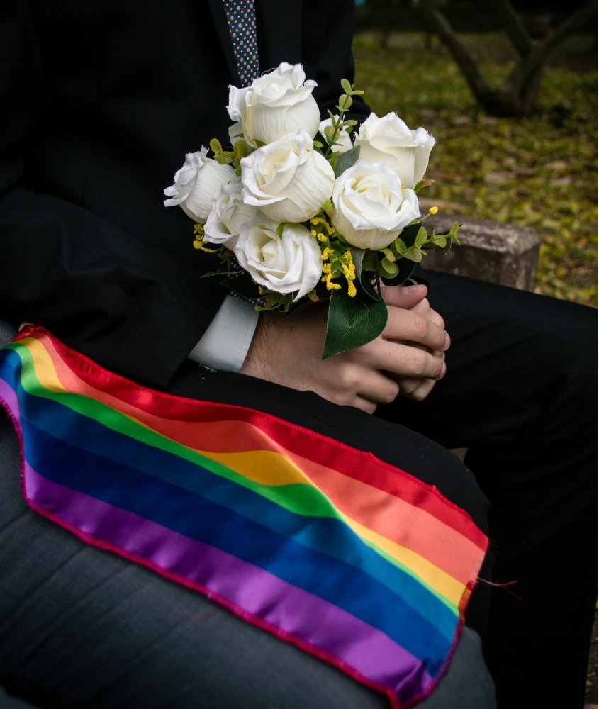 Pessoa segurando um buquê de rosas brancas com uma bandeira LGBTQIA+ no colo, durante um casamento ou cerimônia