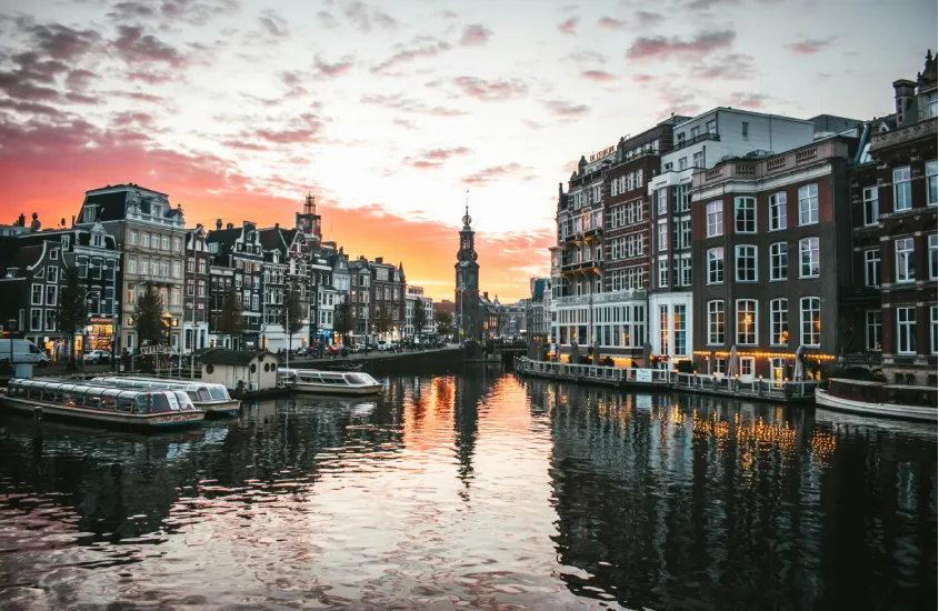 Pôr do sol sobre um canal de Amsterdã, com edifícios típicos refletidos na água e barcos atracados ao longo da margem