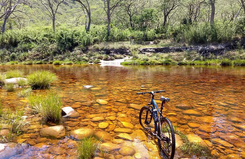 Uma bicicleta parcialmente submersa em um riacho de águas cristalinas com fundo alaranjado de pedras. A paisagem é cercada por árvores e montanhas ao fundo