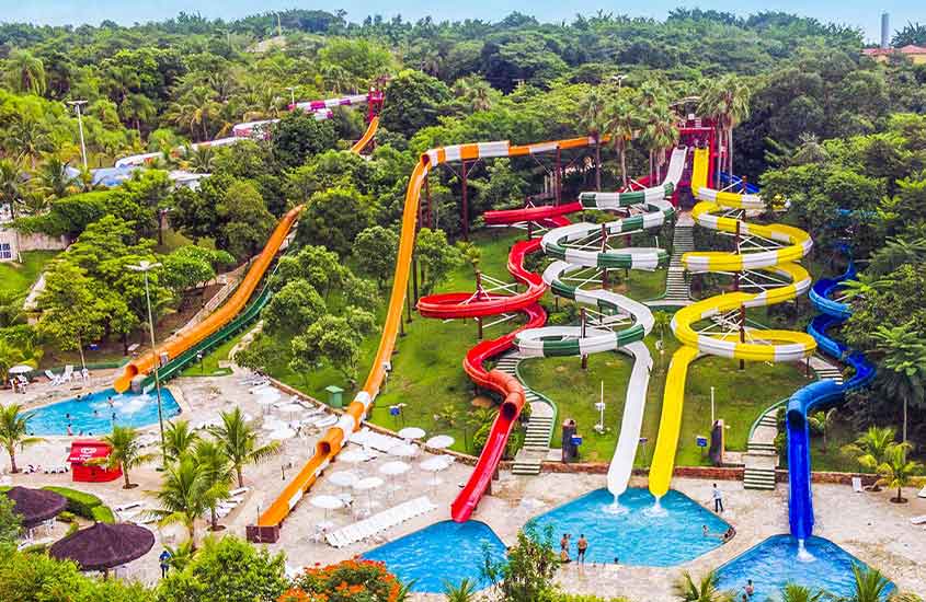 Vista de tobogãs coloridos de um parque aquático rodeado de árvores. Os escorregadores curvilíneos levam a diferentes piscinas, compondo um ambiente de lazer