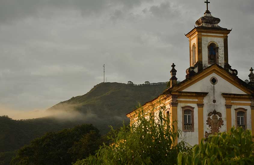 Igreja histórica com fachada colonial em tons de branco e amarelo, com montanhas verdes ao fundo sob um céu nublado