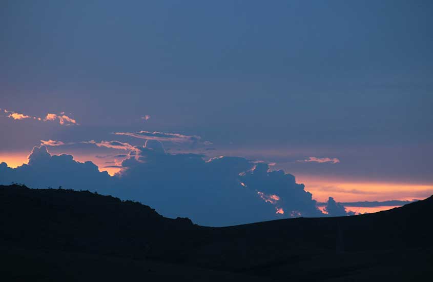 Entardecer com nuvens alaranjadas iluminando o céu, em contraste com as montanhas escuras, capturando um momento sereno e belo da natureza