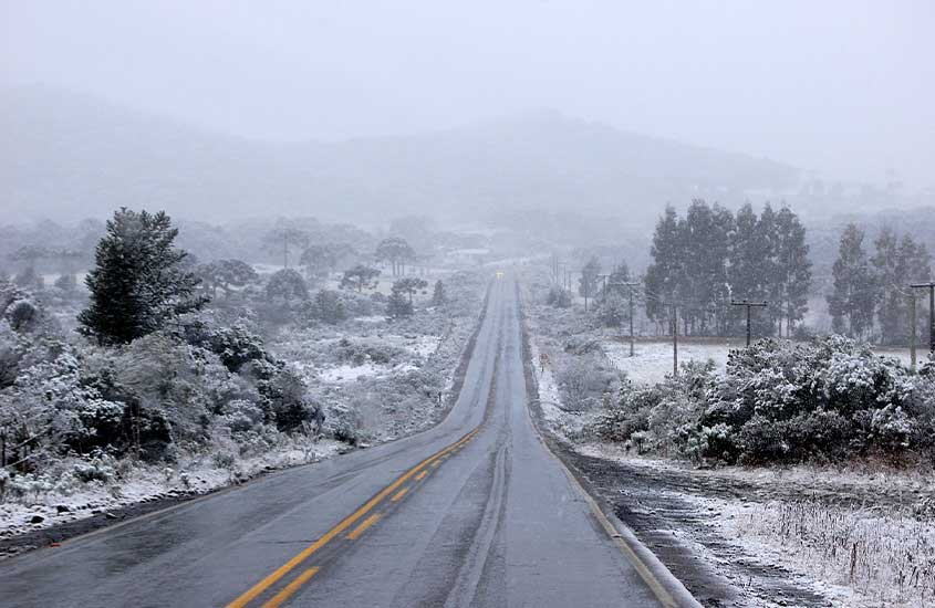 Estrada coberta de neve em São Joaquim, atravessando uma paisagem montanhosa branca e árvores ao longe em meio à neblina