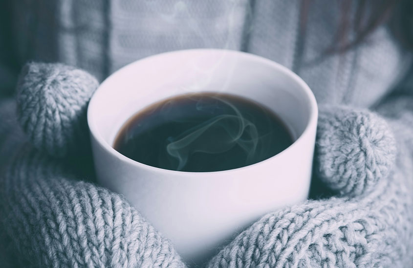 Mãos com luvas de lã cinza segurando uma xícara de café quente, com vapor subindo da bebida