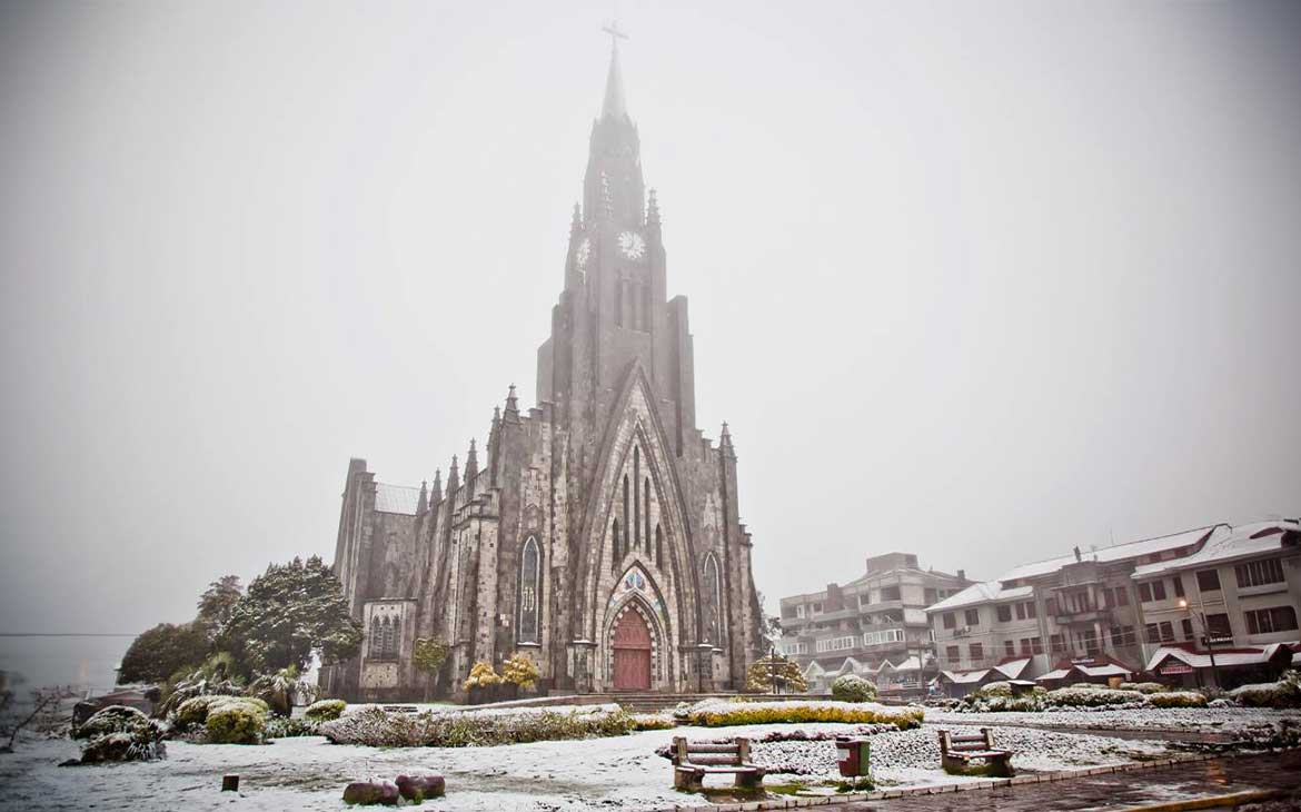 Catedral de Pedra, em Canela, coberta por neve, com bancos e jardins parcialmente cobertos, destacando a atmosfera invernal da cidade