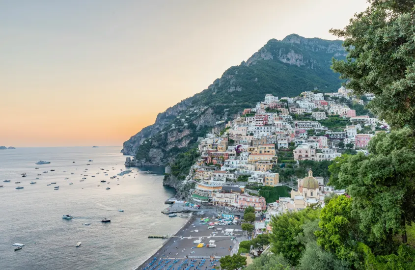 Casas coloridas empilhadas nas encostas de Positano ao entardecer, com barcos flutuando no mar calmo da costa italiana