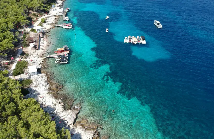 Praia de pedras em Hvar, cercada por vegetação densa e águas azul-turquesa, com barcos e lanchas ancorados