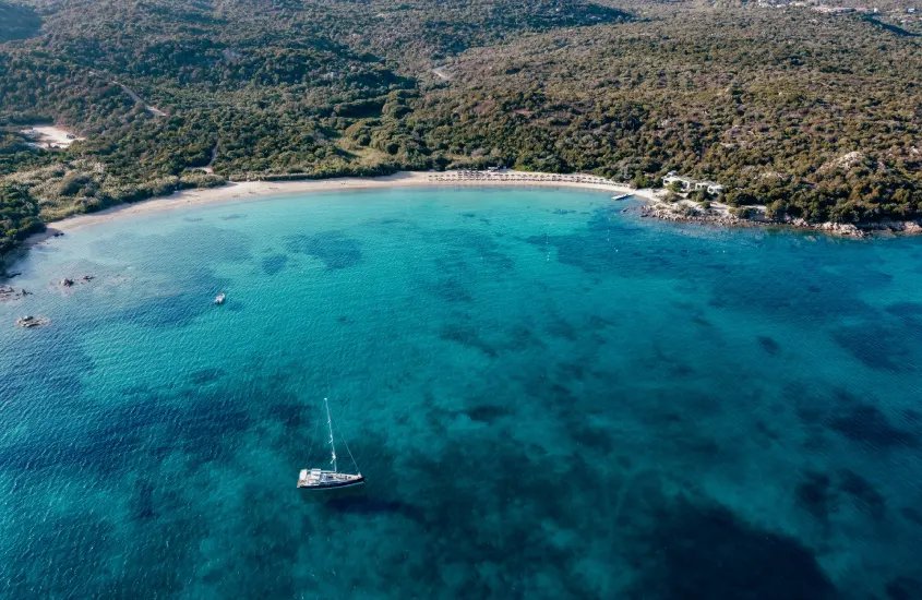 Baía tranquila de águas claras e esmeralda, com um barco à vela ao longe, cercada por colinas verdes na Costa Smeralda