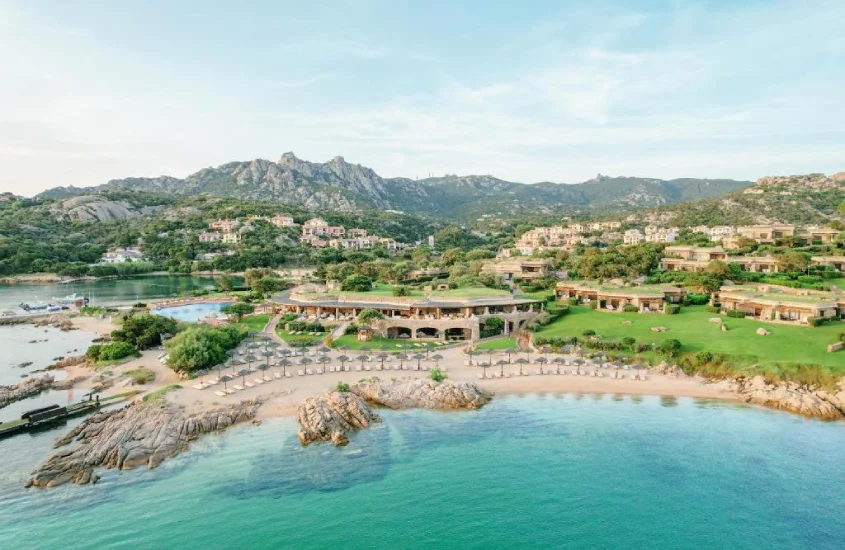 Hotel elegante em Costa Smeralda com praia particular, colinas verdes ao fundo e mar calmo sob o céu azul claro