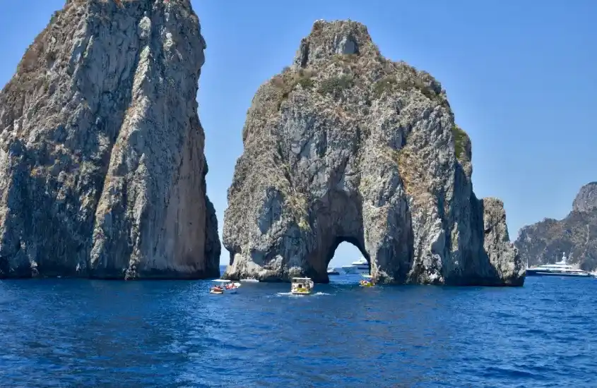 Rochas imponentes dos Faraglioni em Capri, com barcos ao redor e céu limpo, em um típico dia de verão