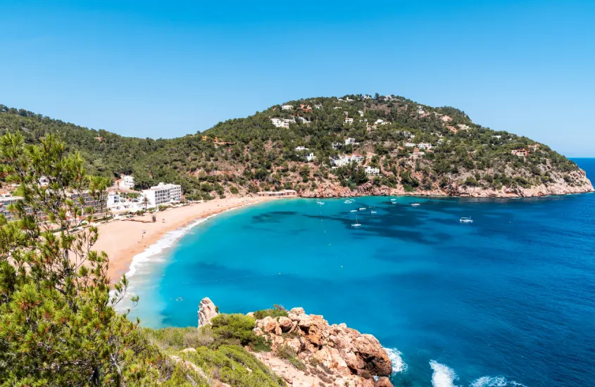 Baía tranquila em Ibiza, cercada por colinas verdes e areia dourada, com o mar azul brilhando sob o céu limpo de verão