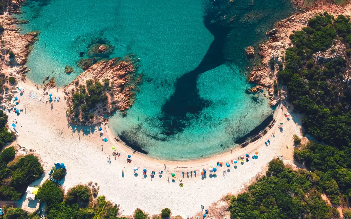 Vista aérea de uma praia com areia branca e águas cristalinas cercadas por rochas e vegetação, com guarda-sóis coloridos espalhados