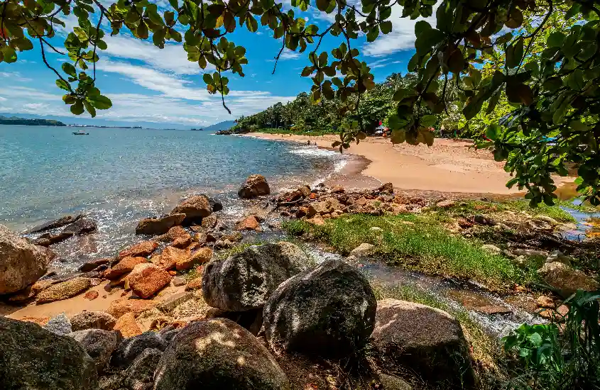 Praia tranquila em Ilhabela ao meio-dia, com rochas e vegetação exuberante enquadrando a paisagem