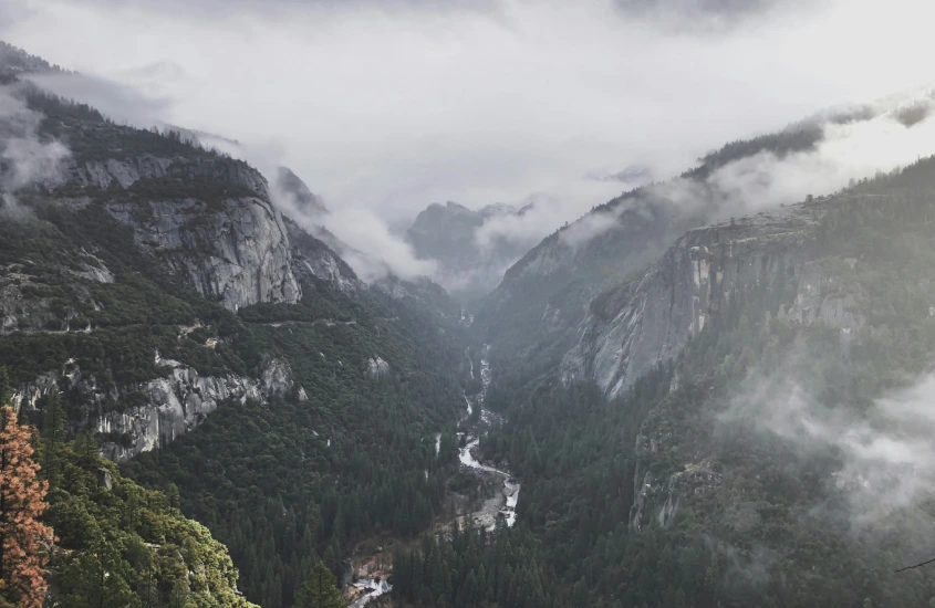 Um vale profundo com montanhas rochosas e árvores escuras cobertas por uma camada de neblina