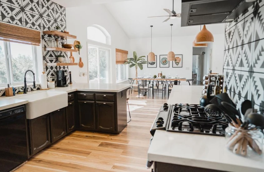 Cozinha moderna e iluminada, decorada com padrões geométricos em preto e branco nas paredes. O ambiente é espaçoso, com uma mesa de jantar ao fundo e detalhes como prateleiras de madeira