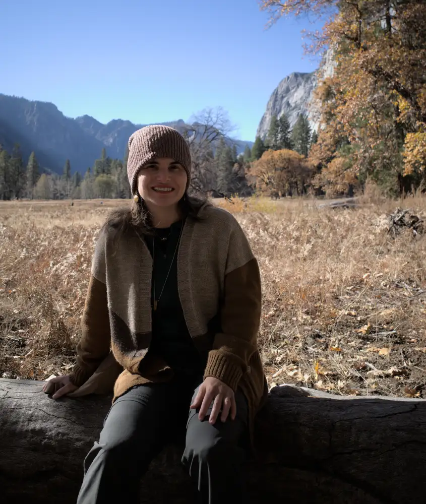 Bárbara Rocha sorridente está sentada sobre um tronco de árvore seco em um campo aberto no Yosemite. Ela usa um gorro de lã e um casaco de malha em tons de marrom. Ao fundo, é possível ver montanhas, árvores e um céu claro e azul.