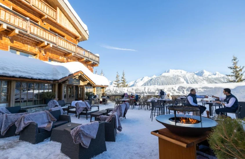 Terraço ao ar livre coberto de neve, com mesas e cadeiras aconchegantes, oferecendo vista para as montanhas alpinas