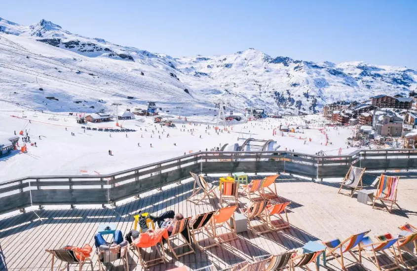 Terraço com cadeiras de praia coloridas, voltado para uma vasta área de esqui em uma montanha nevada