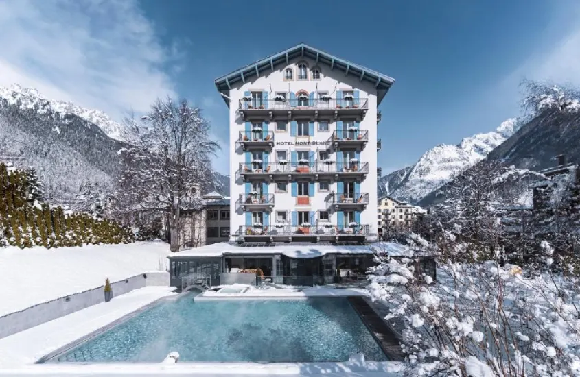 Fachada imponente do Hôtel Mont-Blanc com piscina aquecida ao ar livre e neve ao redor, com montanhas ao fundo