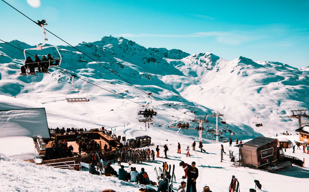 Esquiadores aproveitam as pistas cobertas de neve com teleféricos subindo as montanhas ao fundo, enquanto um deck ao ar livre reúne visitantes no cenário deslumbrante de Val Thorens, nos Alpes franceses.