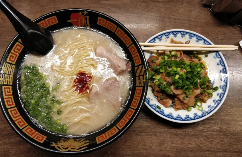 Uma tigela de ramen Ichiran com caldo cremoso, macarrão, cebolinha picada e pedaços de carne suína, acompanhada por um prato de carne de porco com cebolinha e hashis