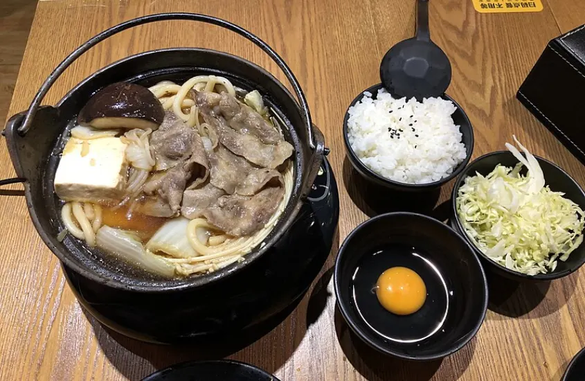 Um prato de sukiyaki quente servido em uma panela, com carne bovina, tofu, cogumelos, macarrão udon e um ovo cru ao lado, acompanhado de arroz e vegetais frescos