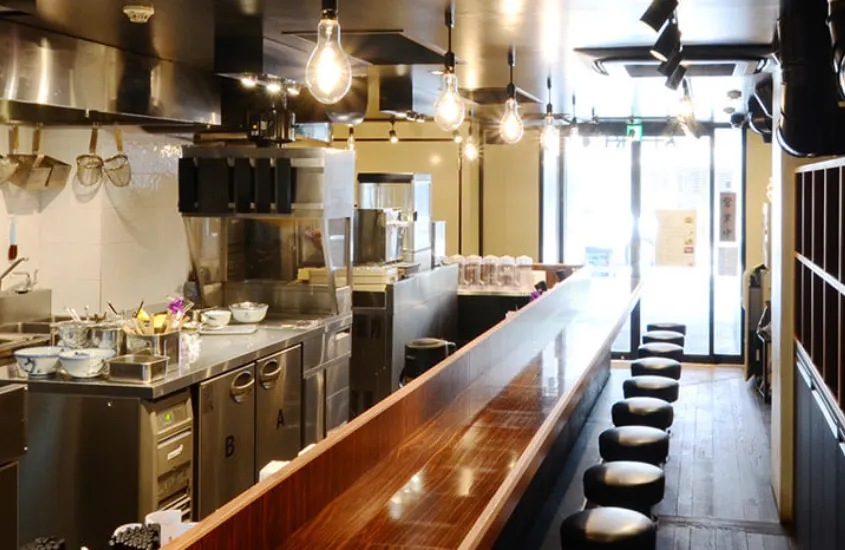 Interior de uma cozinha moderna de um restaurante de ramen no Japão. As panelas e fogões estão à vista, com um longo balcão de madeira e bancos para os clientes