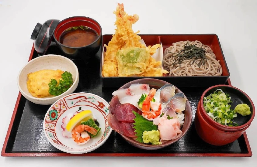 Uma refeição japonesa completa servida em um conjunto de bandejas lacadas. O prato inclui sashimi, tempura crocante, soba (macarrão japonês), arroz e sopa